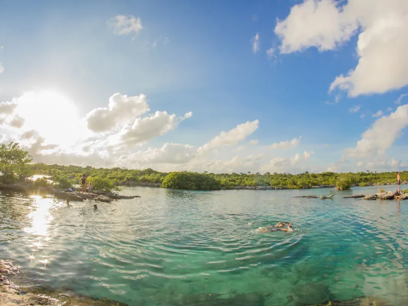 A swimmer in calm lagoon water ringed by low mangroves, with sun and white clouds above