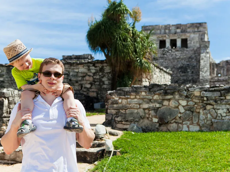 A father carries his young son on his shoulders in front of the stone ruins at Tulum
