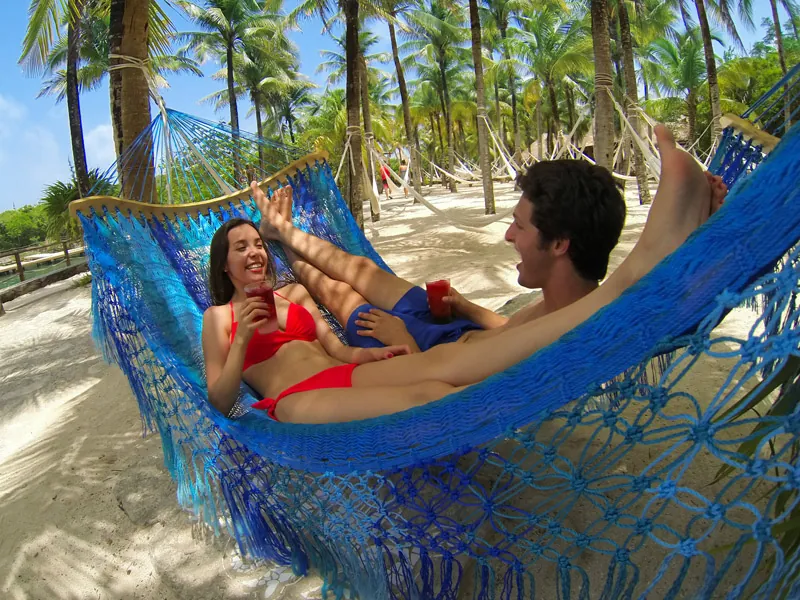 A couple with drinks relax in a blue hammock under palm trees at Xel-Há