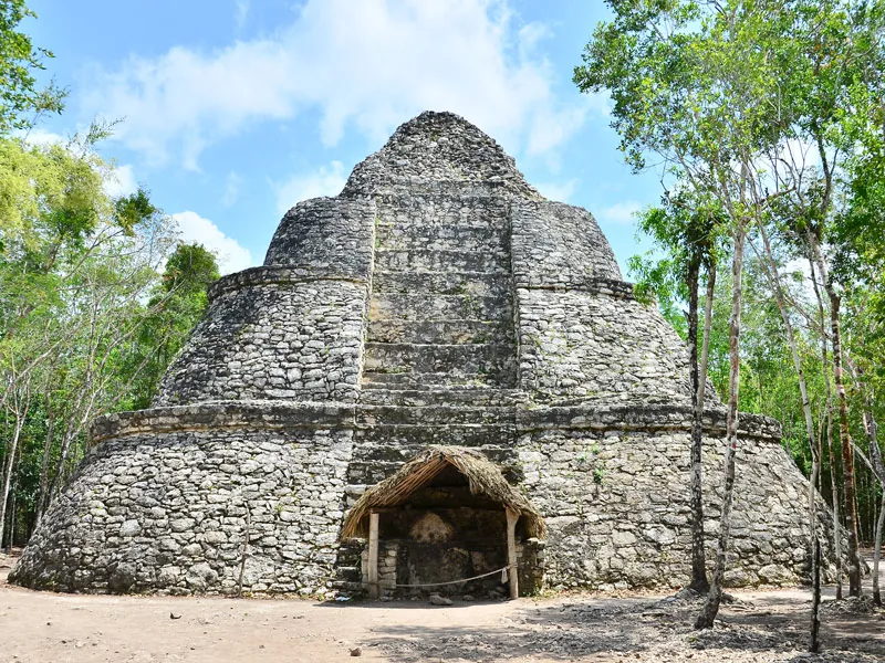 A rounded, terraced stone temple at Cobá with a thatched shelter at its base, framed by trees