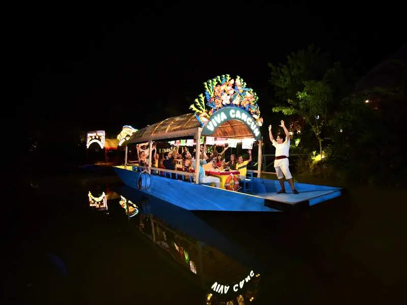 A lit trajinera boat with a flowered Viva Cancún arch glides along a dark canal at night