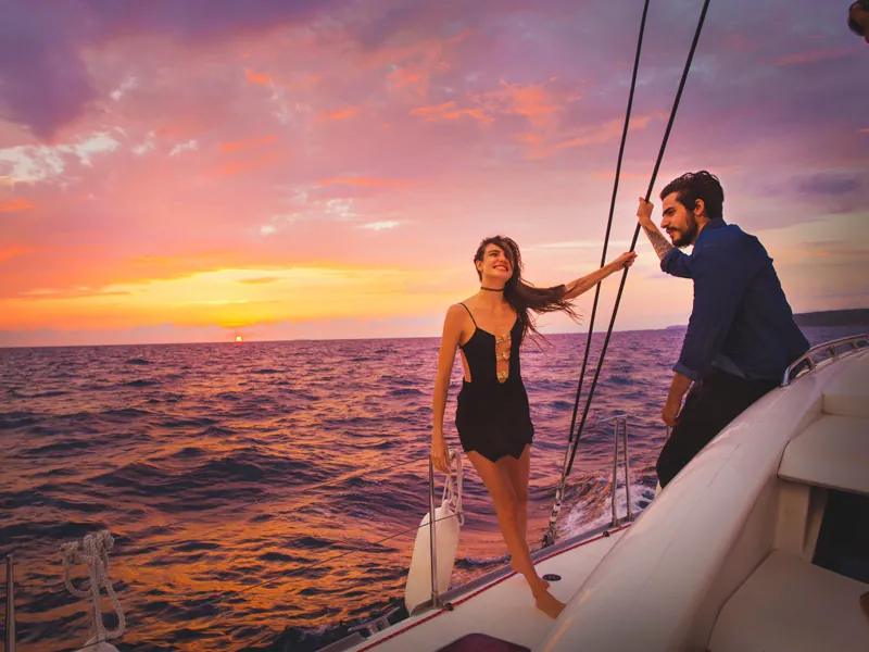 A couple at the bow of a catamaran with the sun setting over the sea behind them