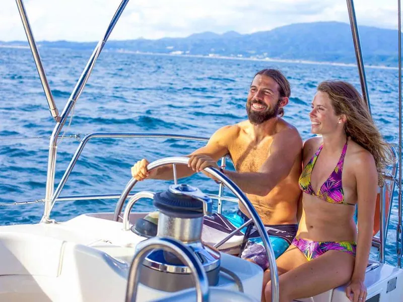 Couple smiling at the helm of a sailboat, green mountains across the water behind