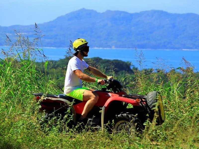 Rider on a red ATV in tall grass with the blue water of Banderas Bay and hills behind, Punta de Mita