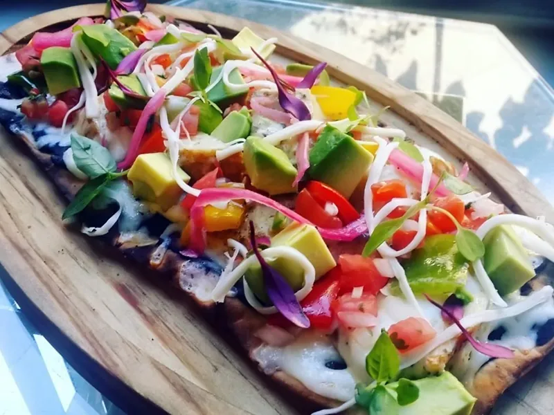 Tostada topped with avocado, tomato, peppers and sprouts on a wooden board