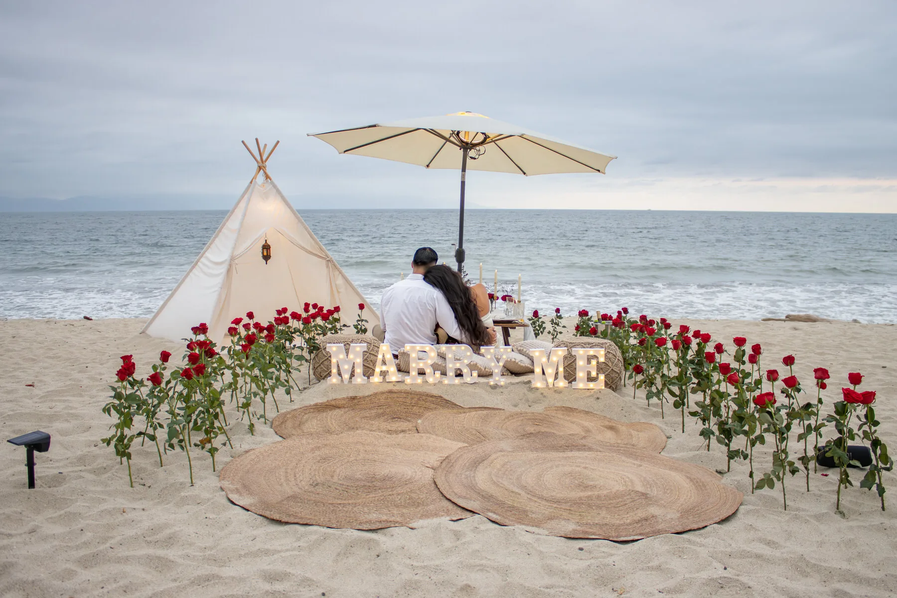 Couple embracing on the beach behind lit MARRY ME letters, red roses, a canvas tipi and a candlelit table facing the sea
