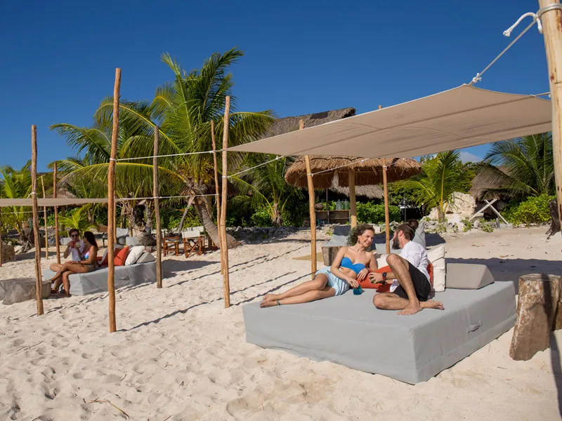 A couple relaxing on a shaded daybed on white sand among palms in Tulum