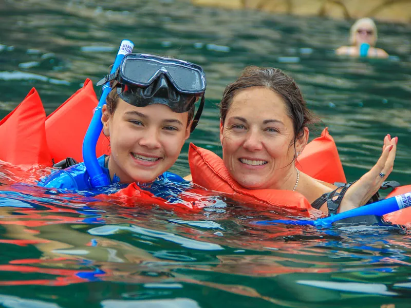 Two snorkelers in orange vests smiling at the surface with masks on their heads