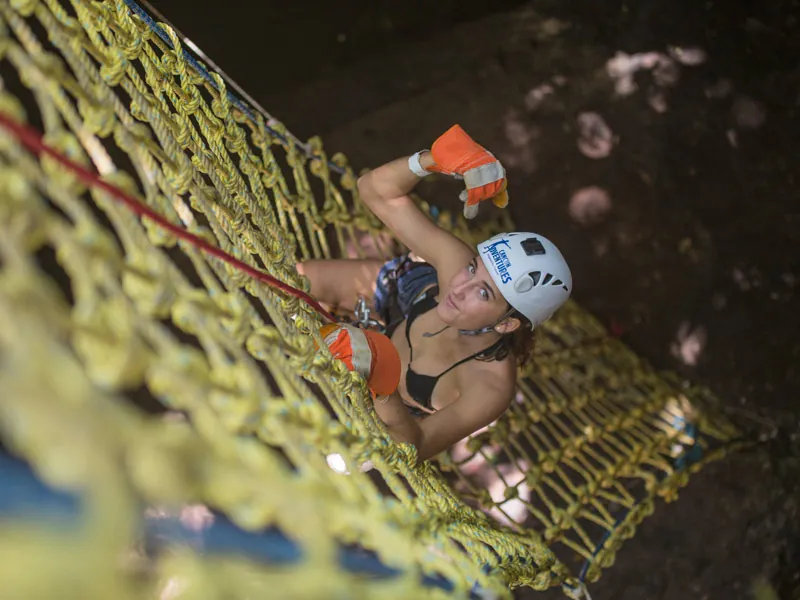 A woman in a helmet and gloves climbing a rope net in a dim cenote