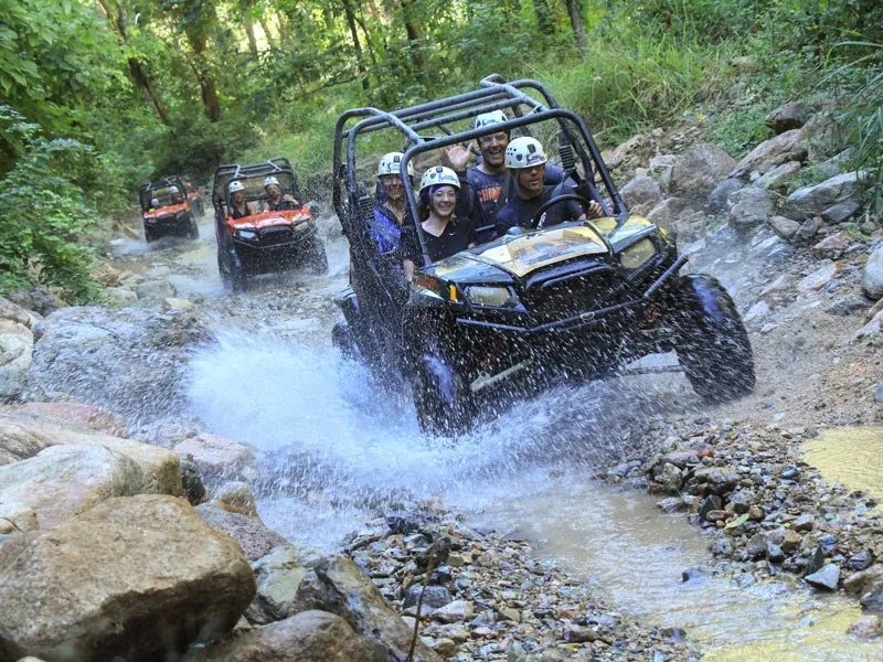 Convoy of all-terrain vehicles splashing through a jungle stream, guests in white helmets