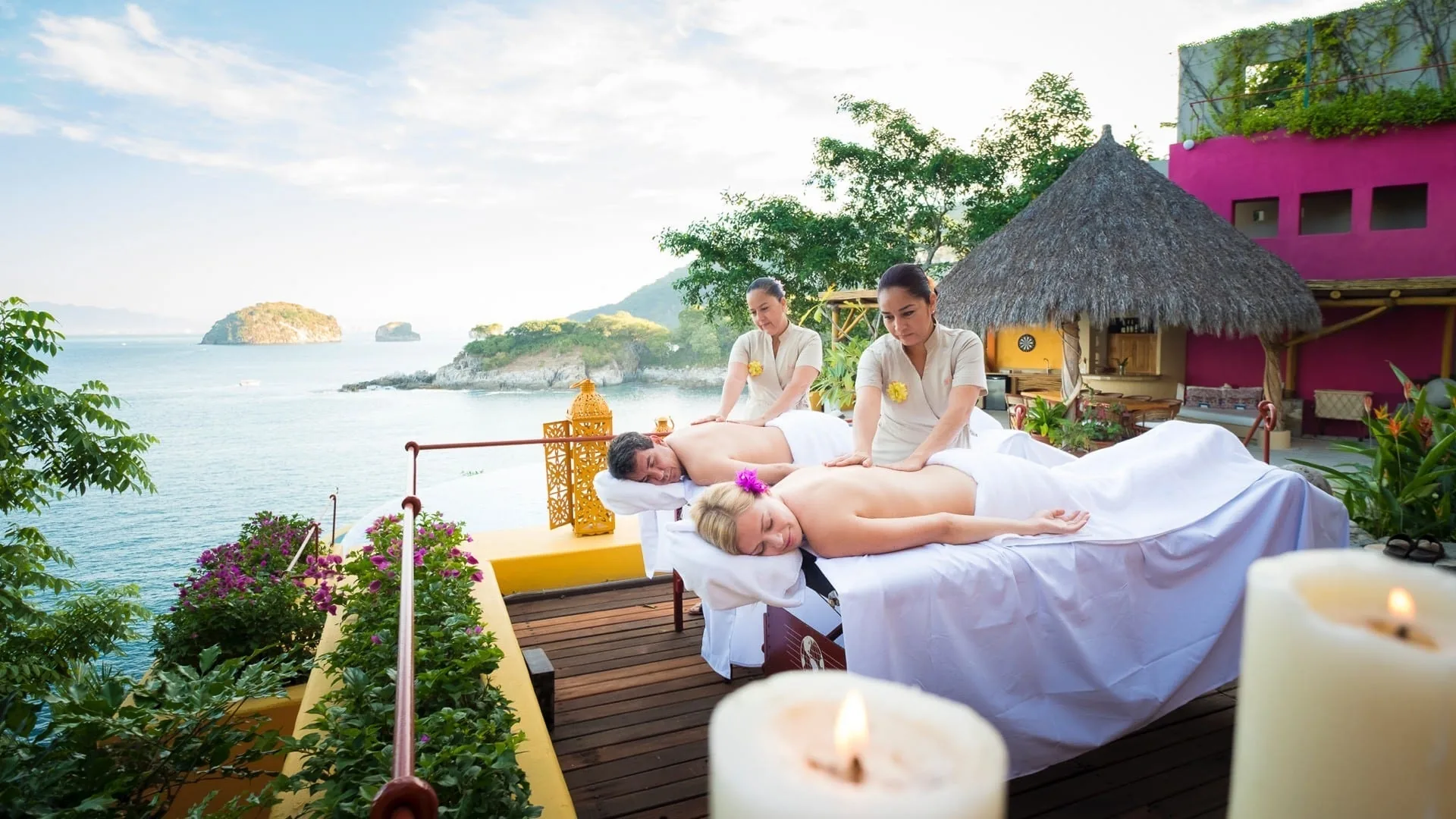 Two therapists giving a couples massage on a candlelit villa deck above the sea and rocky islands of Banderas Bay