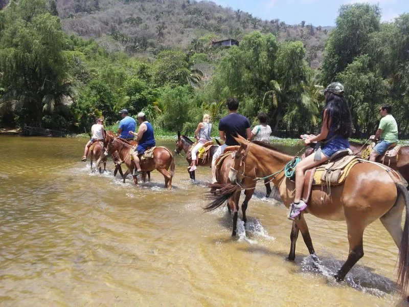 Riders on horseback crossing a shallow river under trees near Quimixto