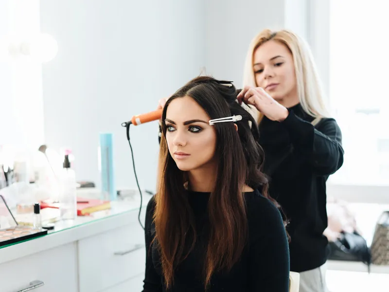 Stylist curling a dark-haired guest’s hair in front of a lit mirror
