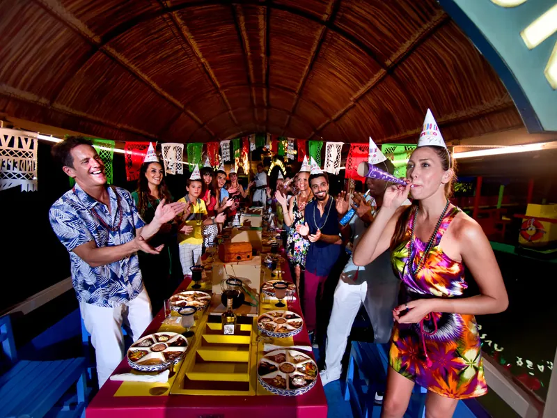 Guests in party hats celebrate at a long dinner table under the roof of a trajinera