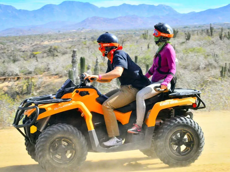 Two riders sharing an ATV among cactus with mountains behind