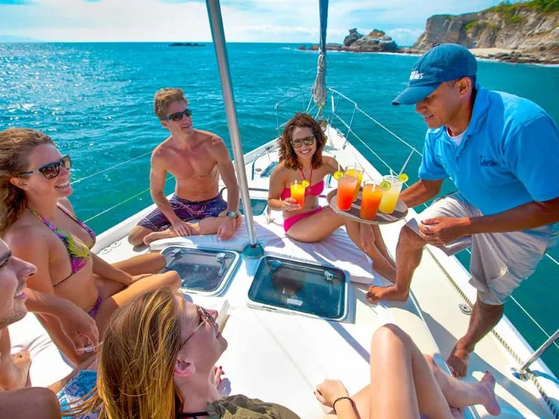 Crew member serving cocktails to guests relaxing on the foredeck near a rocky coast