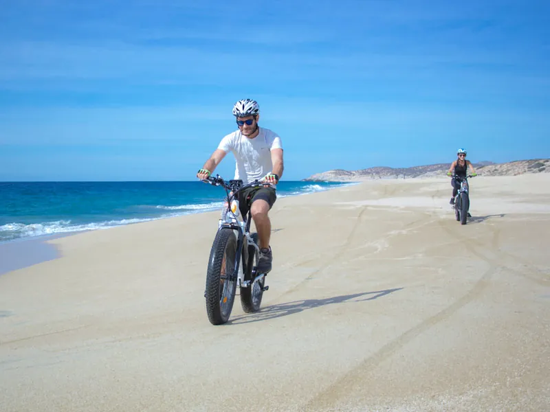 Two riders on electric fat bikes on a wide empty beach beside the Pacific