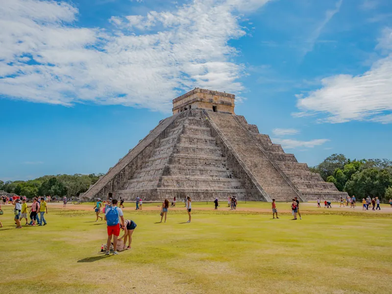 The stepped pyramid of El Castillo at Chichén Itzá under a blue sky, with visitors on the lawn