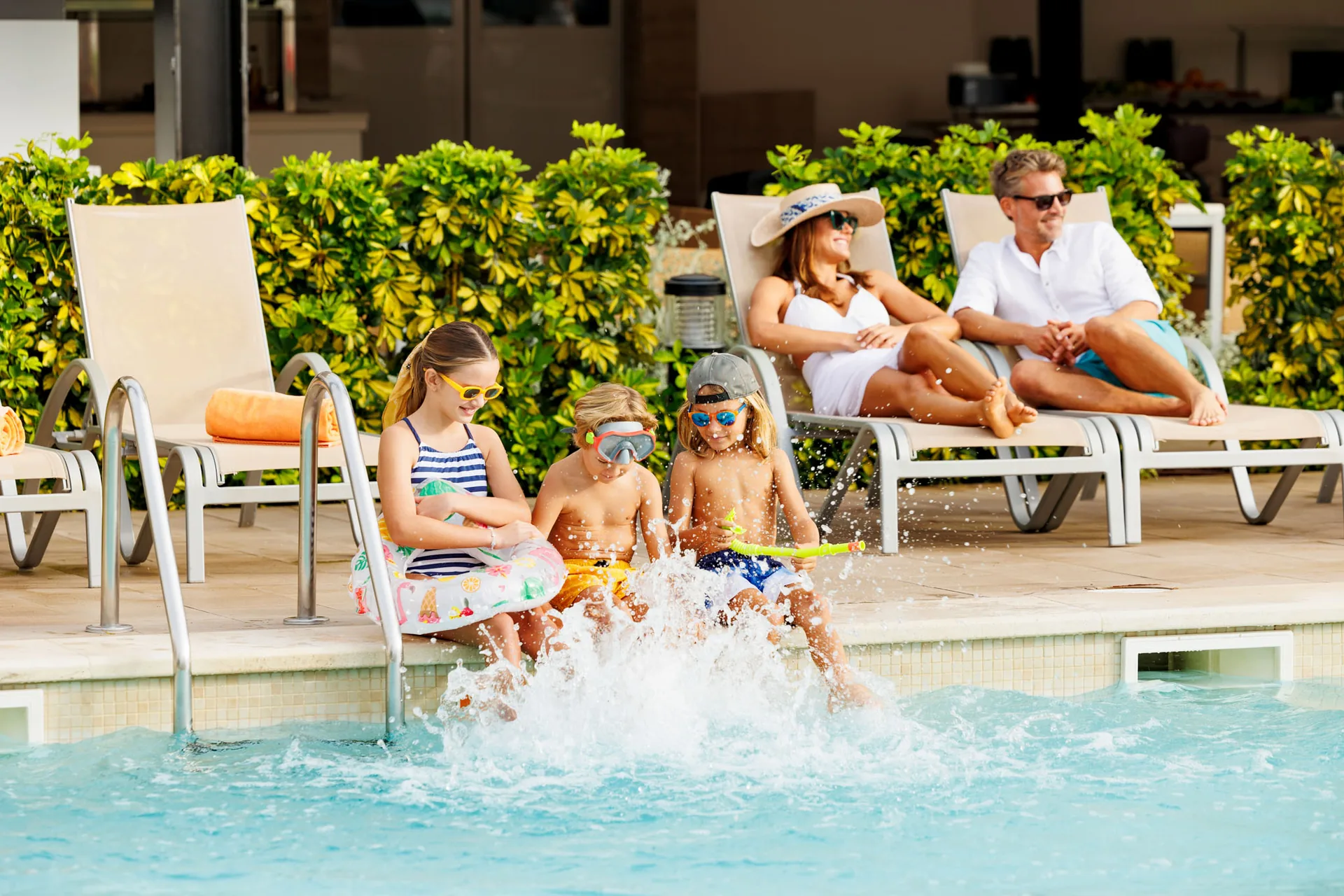 Three children splashing their feet in a pool while their parents relax on loungers behind them