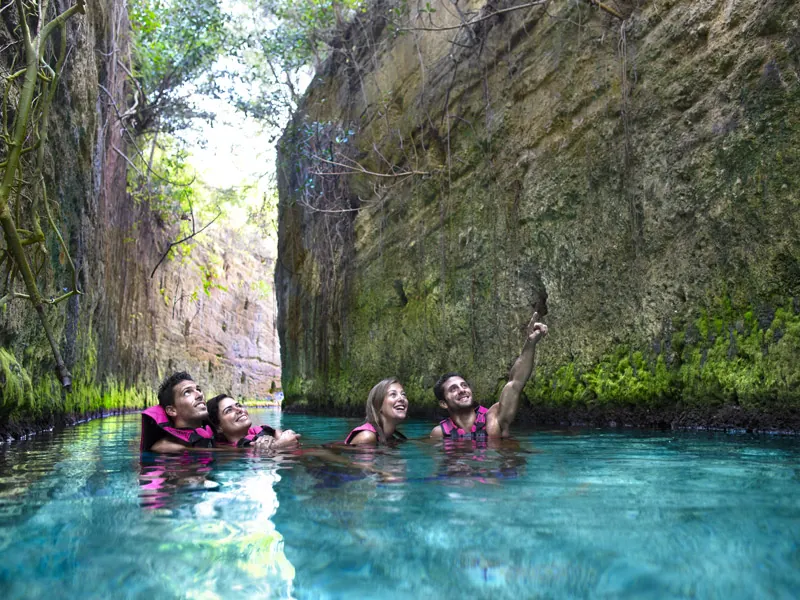 Four swimmers in life vests float through an underground river between tall rock walls at Xcaret