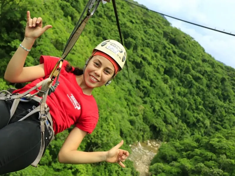 Smiling guest in a helmet and harness on a zip line high above a green river valley at Canopy River