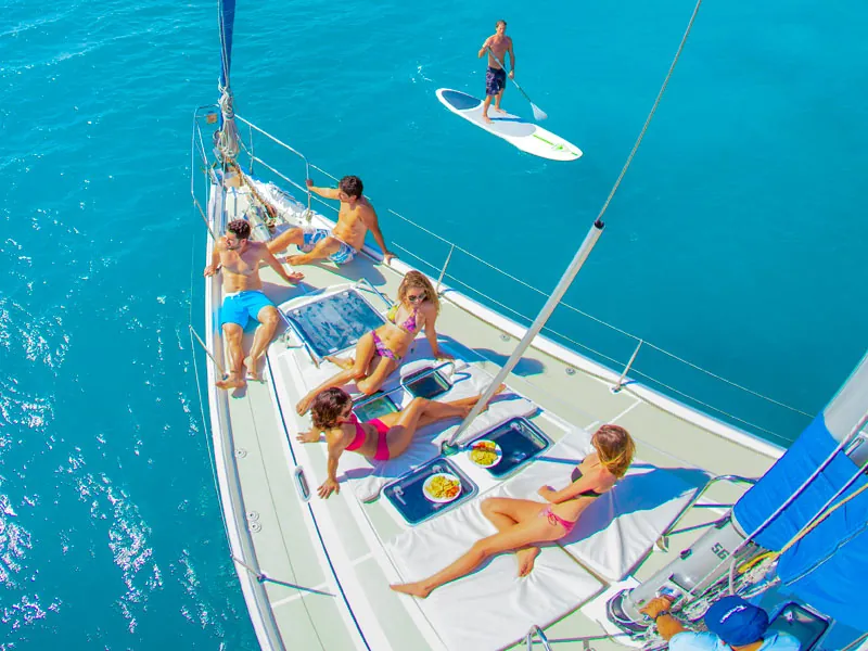 Guests sunbathing on a sailboat foredeck while one paddleboards alongside in turquoise water