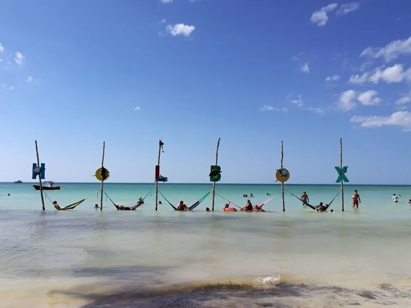 A row of hammocks strung on posts in shallow turquoise water off Holbox, with swimmers resting
