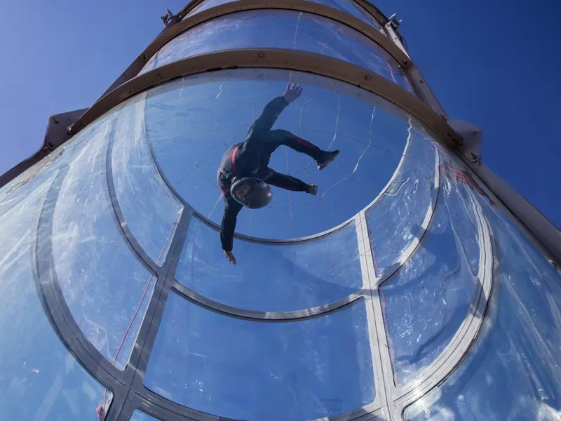 Flyer floating inside a glass skydiving wind tunnel seen from below against a blue sky