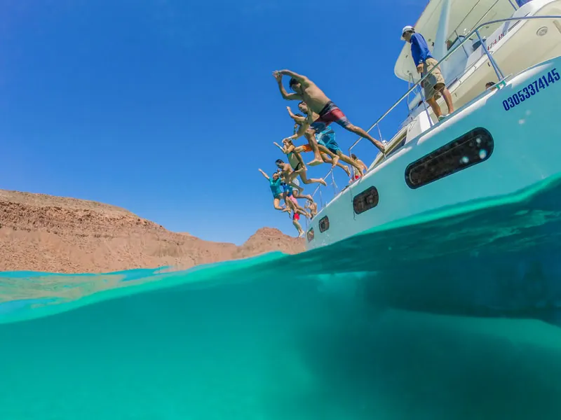 Guests jumping from a catamaran into turquoise water below red cliffs