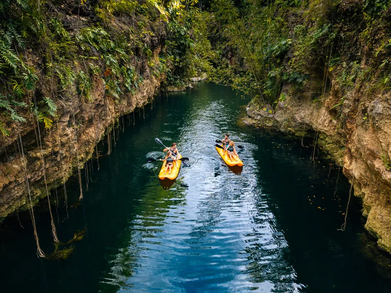 Two kayakers paddle a narrow green-water cenote between steep rock walls hung with roots