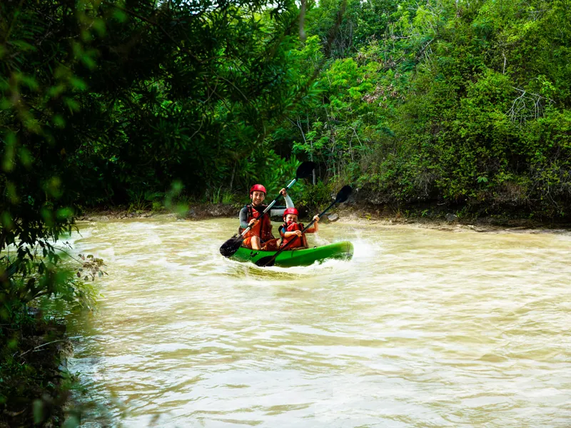 Two paddlers in helmets steer a kayak down a fast brown river lined with jungle