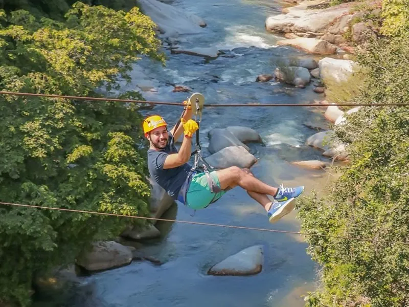 Man in a yellow helmet on a zip line strung low over a boulder-strewn river