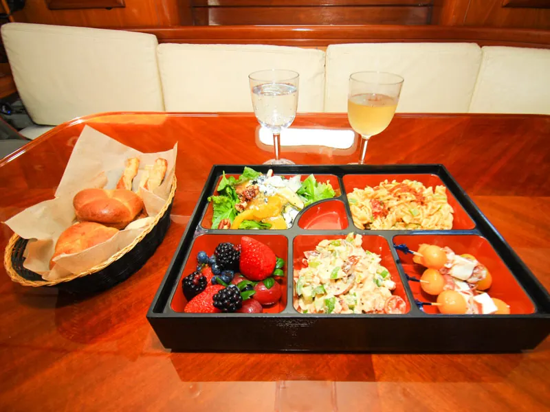 Lunch tray of salads, fruit and bread rolls with two drinks on the sailboat saloon table