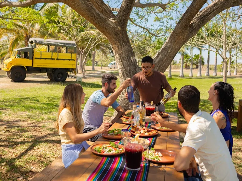 Guests toasting at a long lunch table under a shade tree, the yellow Unimog parked behind