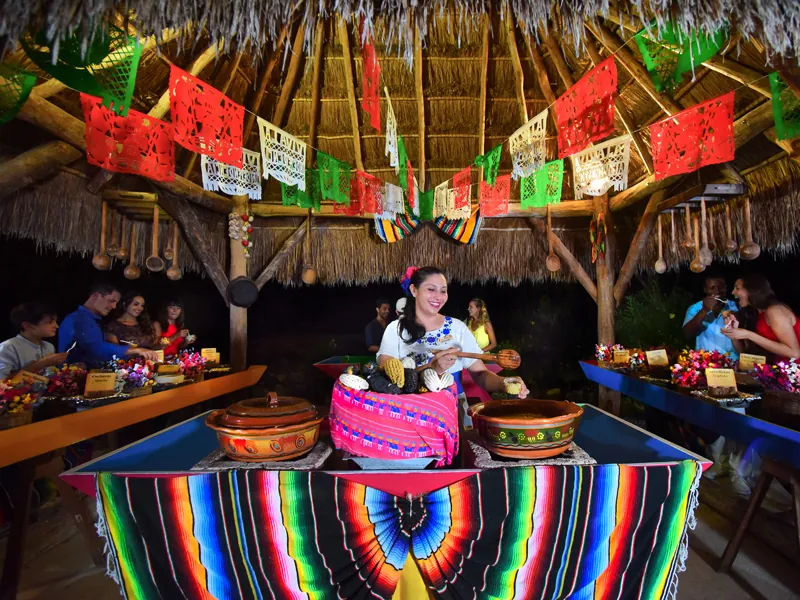 A cook in traditional dress serves from clay pots beneath papel picado banners