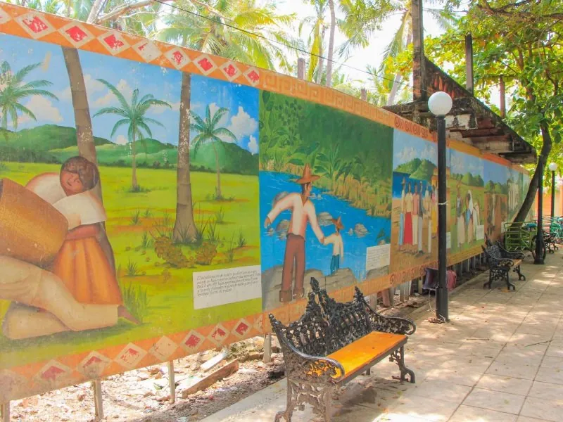 Painted mural panels along a palm-shaded walkway with an iron bench in San Pancho