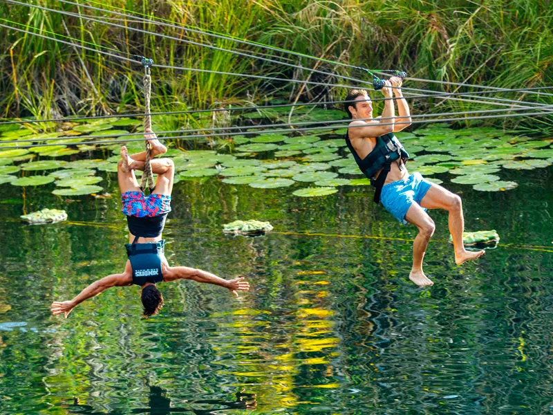 Two guests on zip-lines above an open cenote covered in water lilies, one hanging upside down