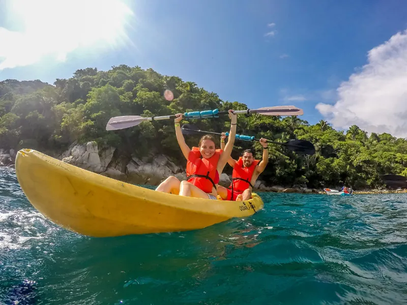 Two kayakers raising their paddles in a yellow kayak near a rocky shore