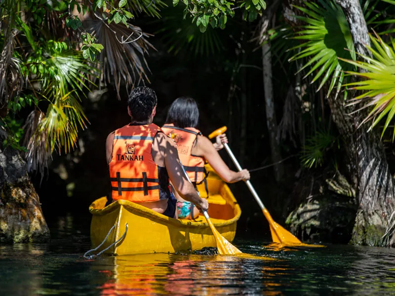 Two paddlers in orange Tankah life vests in a yellow canoe among jungle palms