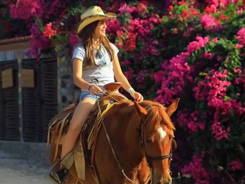 Woman in a straw hat riding a chestnut horse past pink bougainvillea in Higuera Blanca