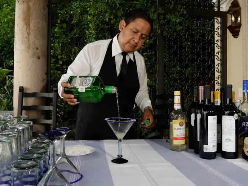 Waiter pouring a margarita at an outdoor bar lined with bottles and glassware