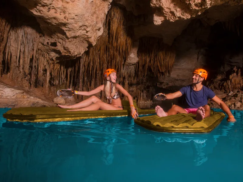 Two guests paddle small rafts by hand through a cave lined with stalactites