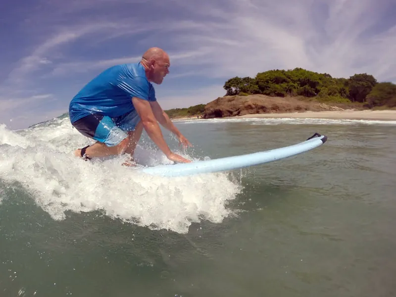 Man in a blue rash guard crouched on a surfboard riding a small wave toward shore