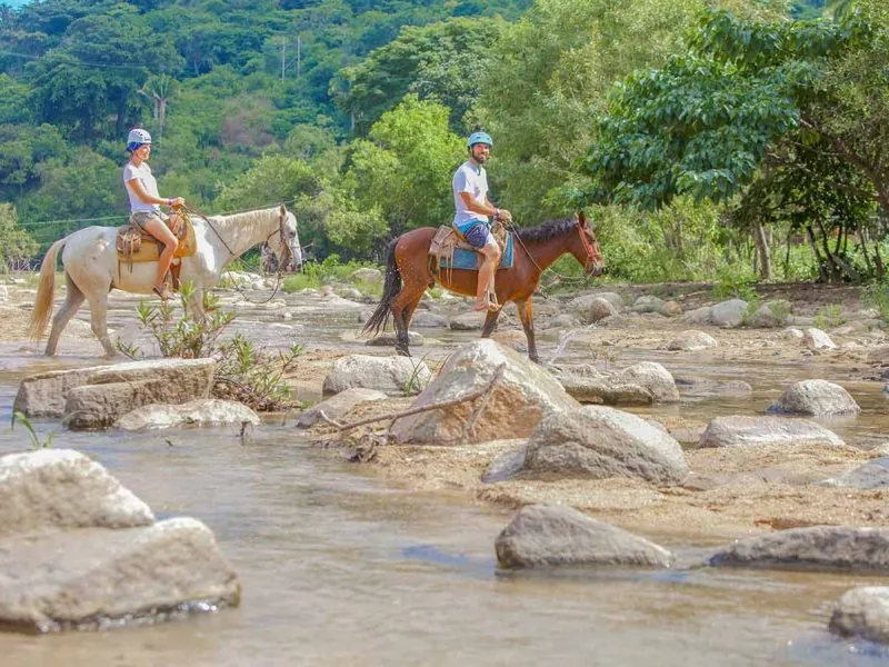 Two riders on horseback crossing a rocky river on the trail to the waterfall