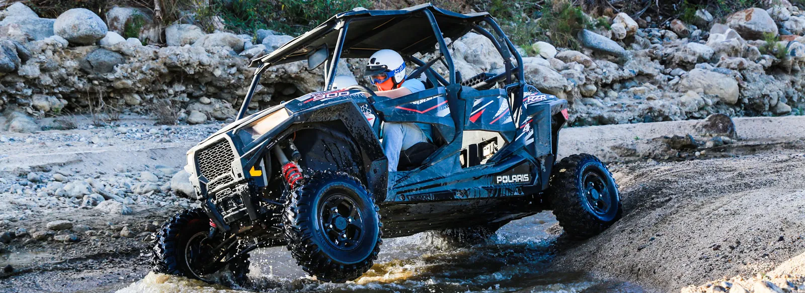 Polaris RZR climbing out of a stream over rocks and sand
