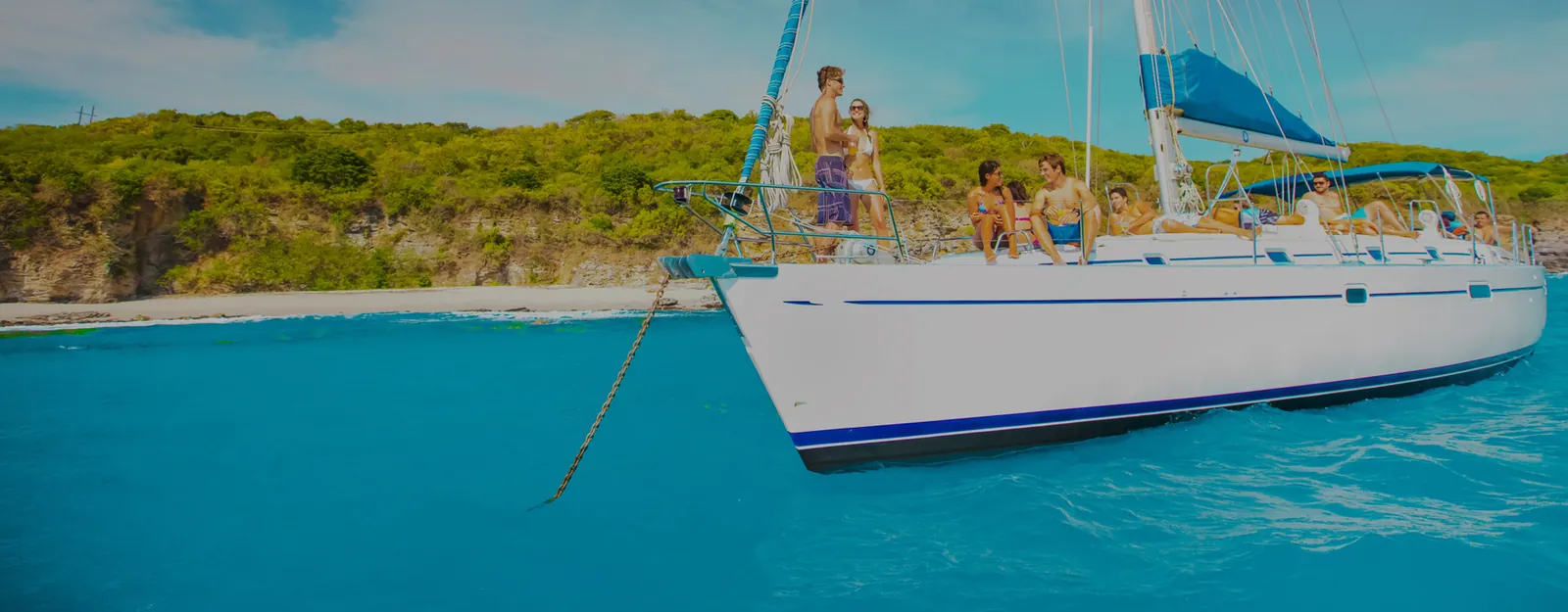 White sailboat anchored off a green shoreline in turquoise water, guests on the foredeck