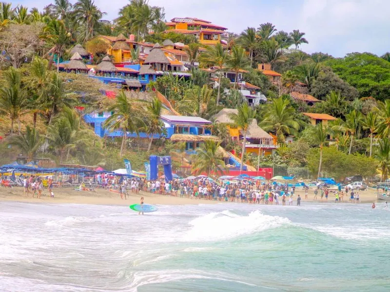 Busy beach at Sayulita with surfers in the waves and colorful houses on the hillside behind