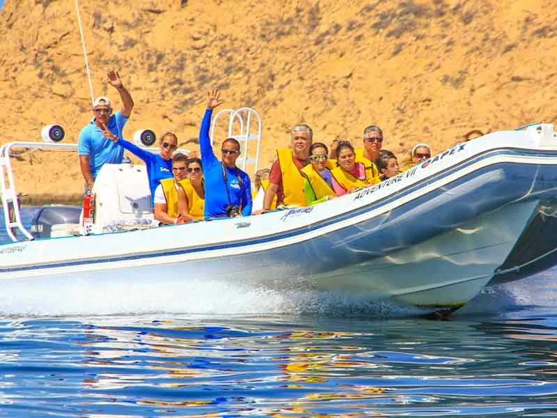 Guests in life vests waving from an inflatable boat below sandy cliffs
