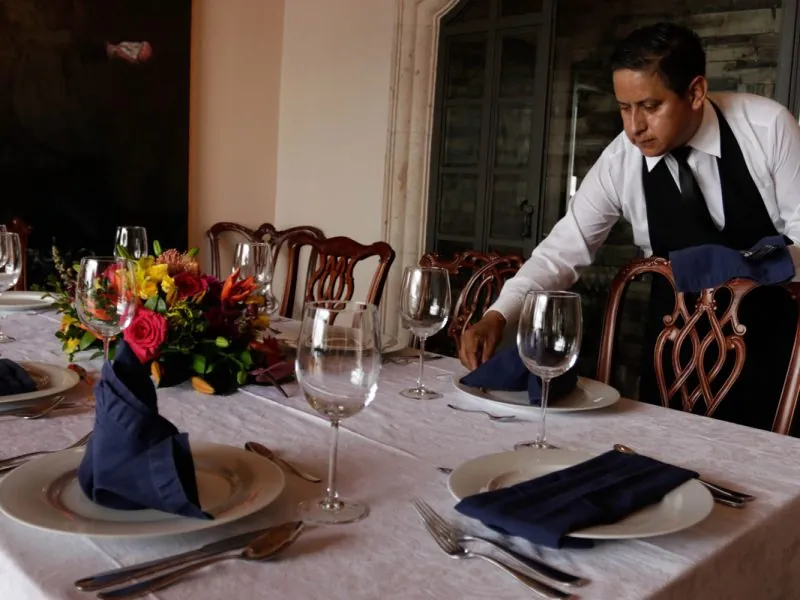Waiter laying place settings on a long white-clothed dining table with flowers