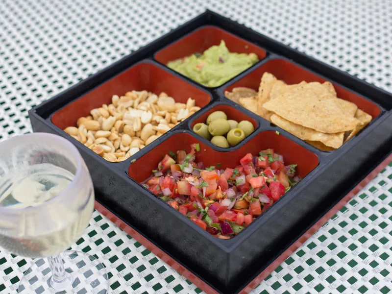 A tray of guacamole, salsa, chips, olives and peanuts beside a glass of white wine